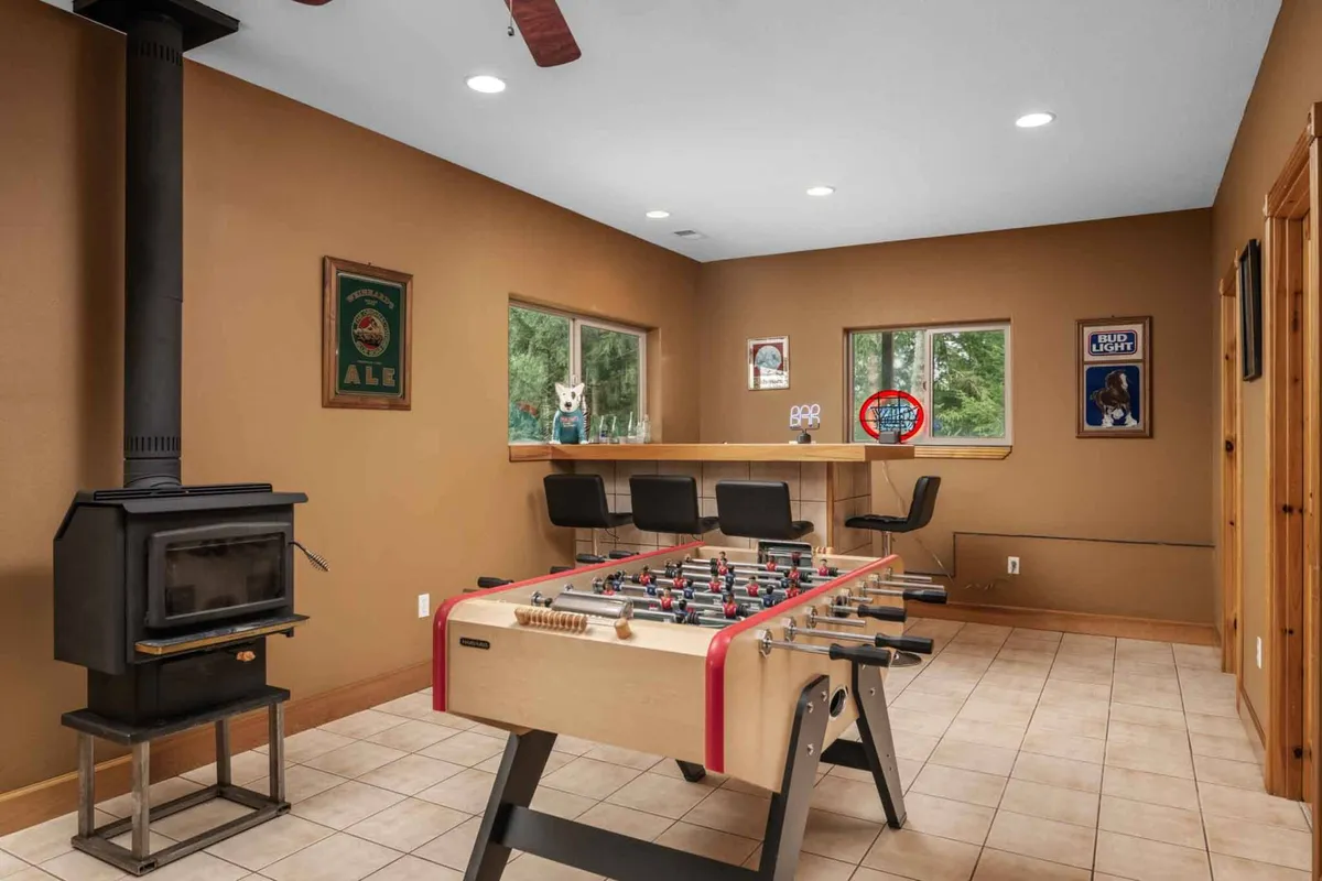 A cozy corner of the entertainment area, featuring a foosball table, a warm wood stove, and a stylish bar area with stools. Large windows provide a view of the greenery outside, infusing the space with a welcoming, friendly mood.