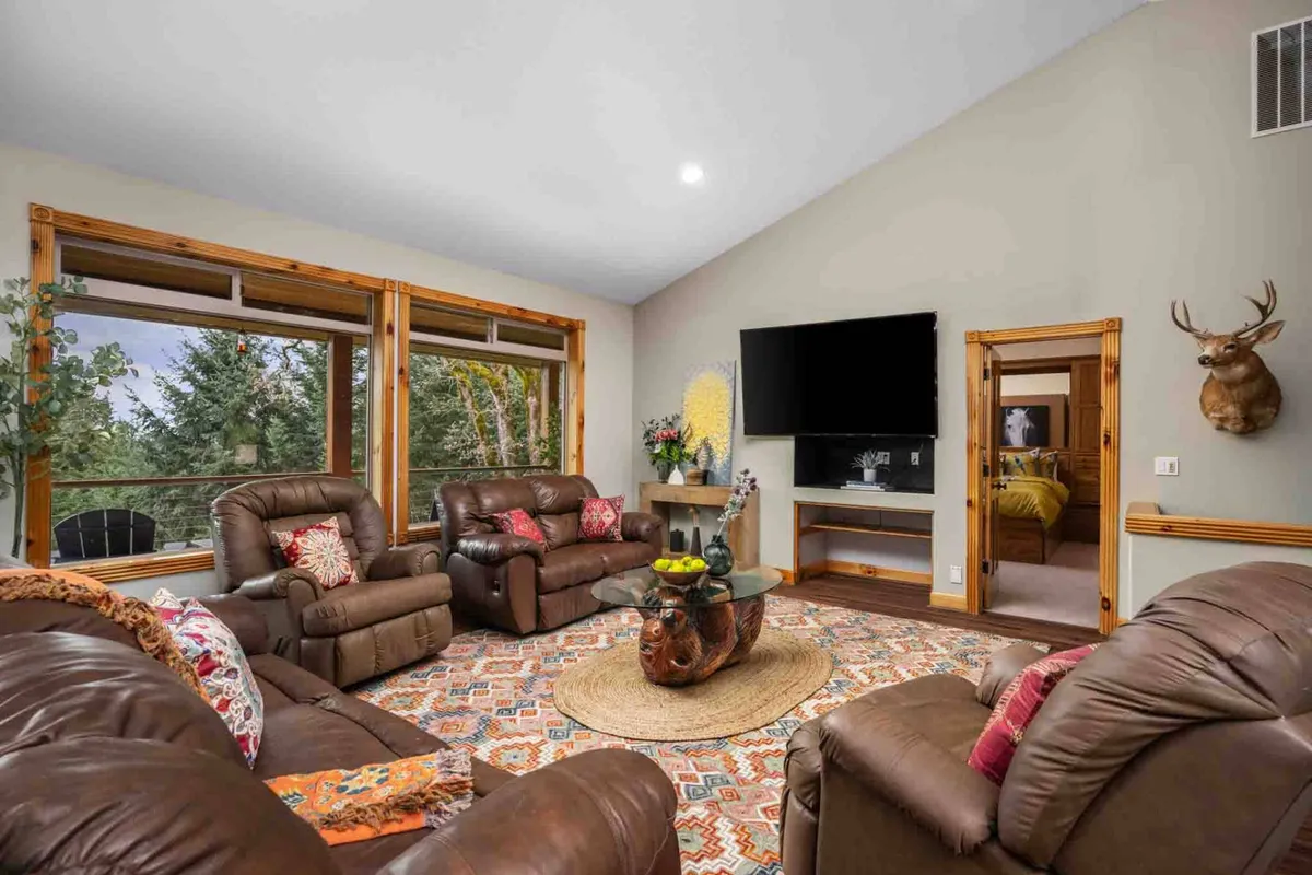 A cozy living area with plush leather sofas arranged for conversation, a decorative rug, large windows offering scenic views, and antler wall décor that adds rustic charm. The TV mounted on the wall invites movie nights and shared entertainment.