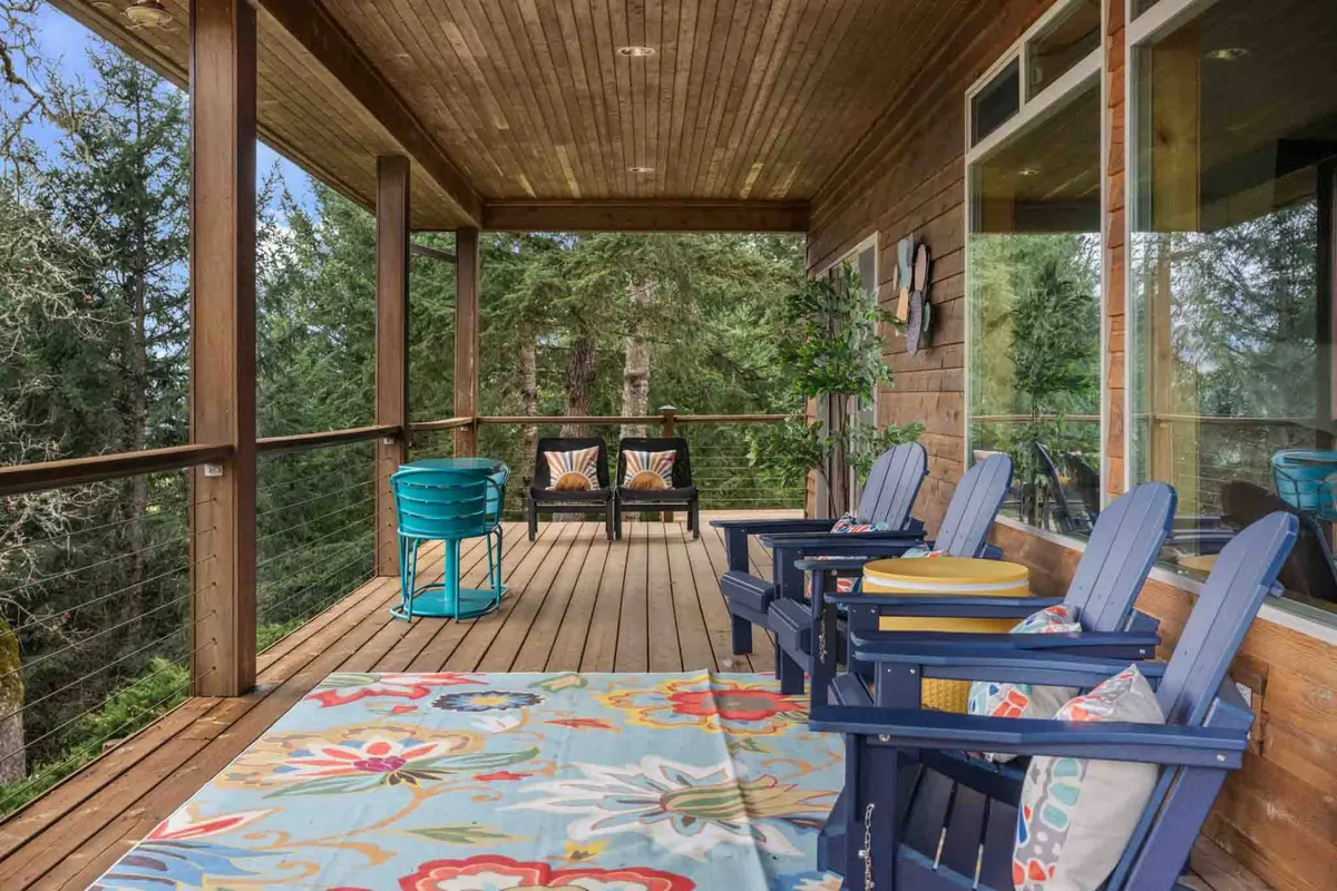A cheerful deck scene with colorful Adirondack chairs and cushions arranged on a bright rug, potted plants adding greenery, and expansive views of trees beyond the railing. The arrangement invites relaxation, morning coffee, or friendly conversations while soaking in the natural beauty.