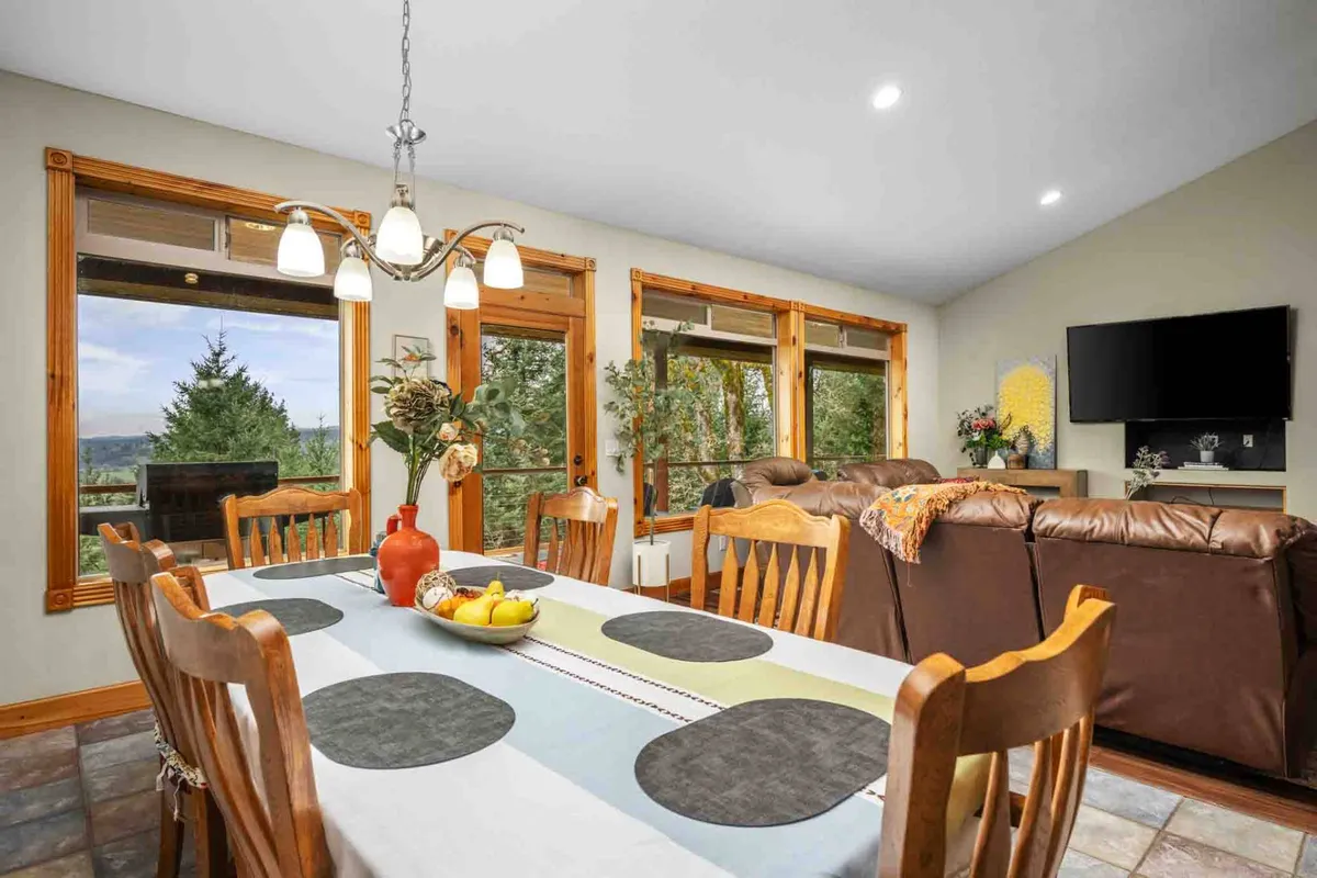 A large, inviting dining table situated near expansive windows with beautiful valley views, seamlessly connected to a cozy sofa seating area. The space feels open, sunlit, and ideal for group meals or celebrations, with details that encourage guests to linger and connect.