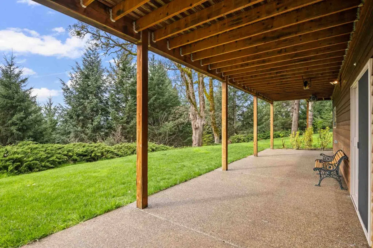 An inviting outdoor seating area beneath wooden beams, with a bench overlooking a grassy lawn and trees beyond. The space is bathed in natural light and surrounded by peaceful greenery, perfect for relaxing or enjoying a picnic.