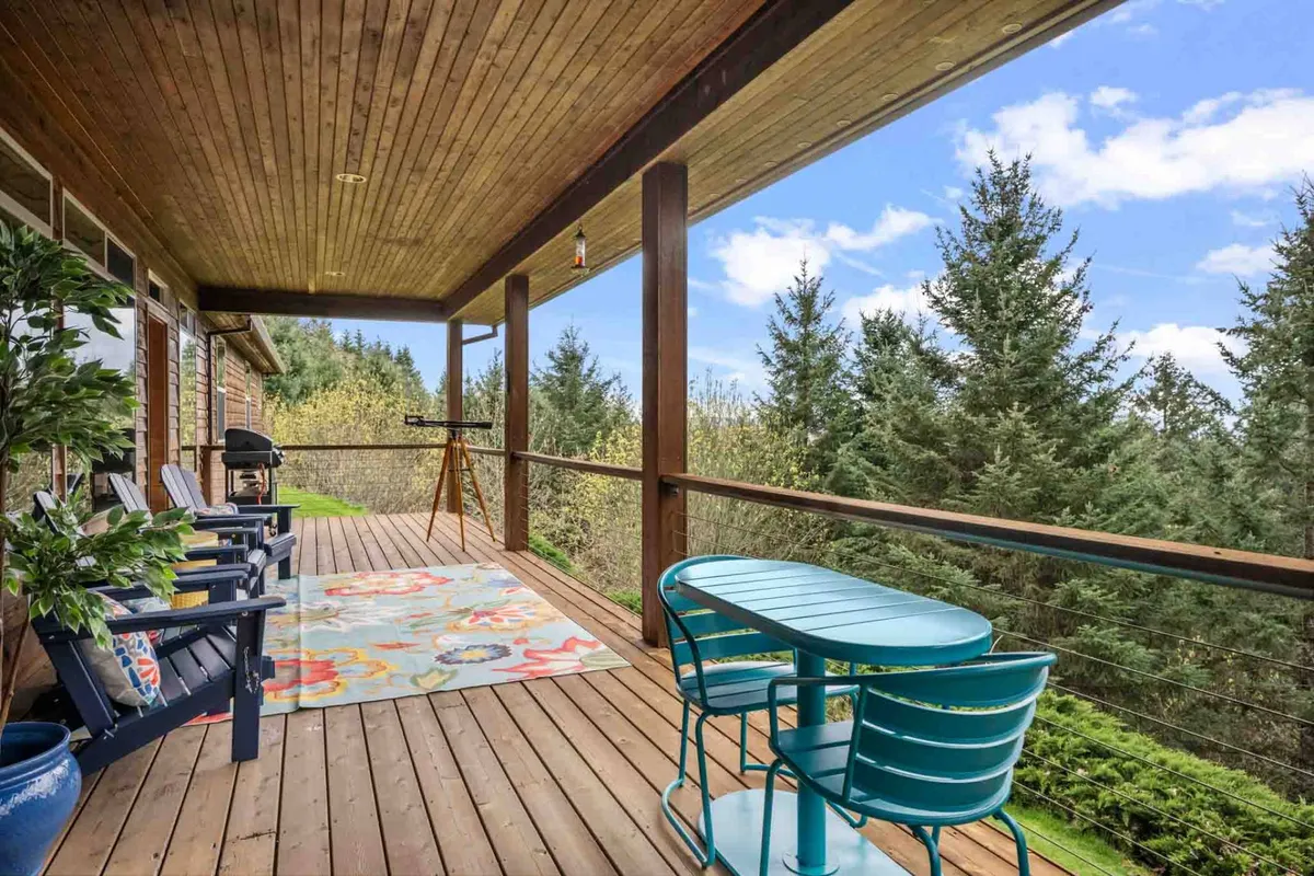 A cozy exterior deck space featuring two inviting chairs and a small table atop a decorative rug, surrounded by lush trees. The view extends out into the greenery, creating a sense of privacy and tranquility—ideal for relaxing with a glass of local wine while soaking in sunset vistas.