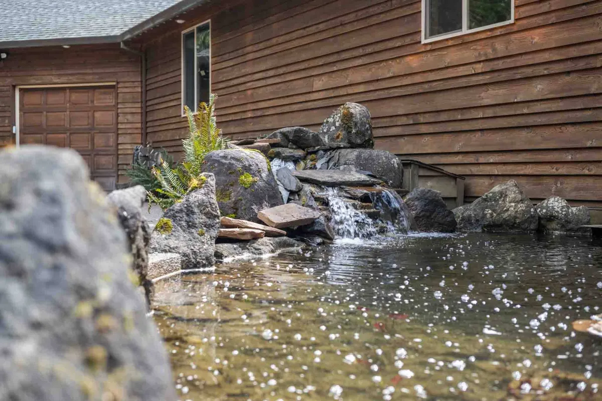 The scene showcases a tranquil water feature with natural rock formations beside the wooden exterior of the house. Lush greenery frames the area, evoking a sense of peace and connection to nature.