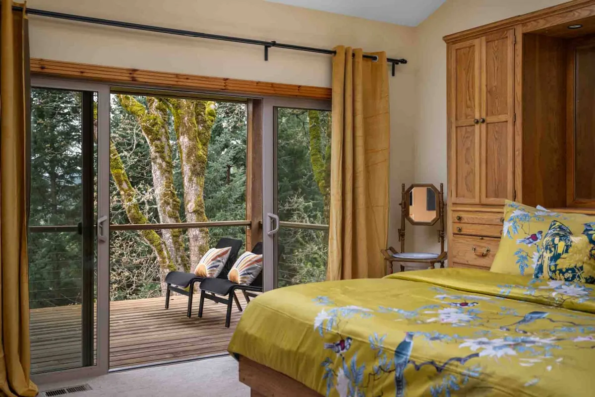 A serene king bedroom with yellow floral bedding, a large sliding glass door, and wooden furnishings. The room opens to an outdoor seating area, with tranquil green views visible through the glass. The soft morning light and natural tones create a sense of calm.
