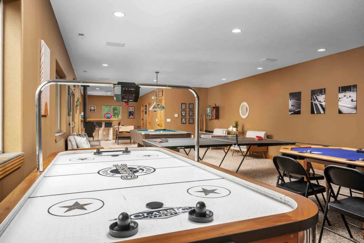 A spacious game room featuring an air hockey table, pool table, and poker table. The room is accented with vibrant artwork on the walls, a comfortable seating area for socializing, and ample space for multiple games to be played at once.