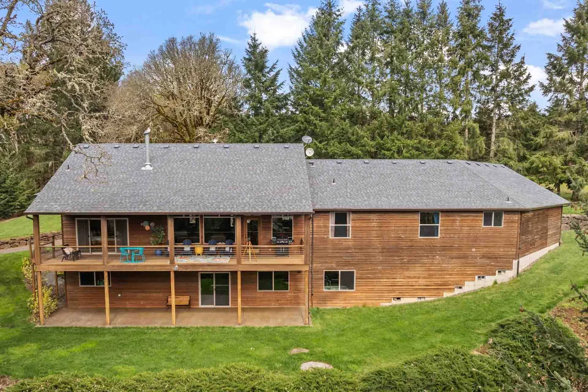 A wide, welcoming wooden deck extends from the main house, bordered by lush greenery and trees. Comfortable seating is arranged for gatherings, with a BBQ grill visible, and large windows connect the indoor living spaces directly to the deck. The expansive valley and natural surroundings are showcased in the background, emphasizing a seamless transition from inside to outside.