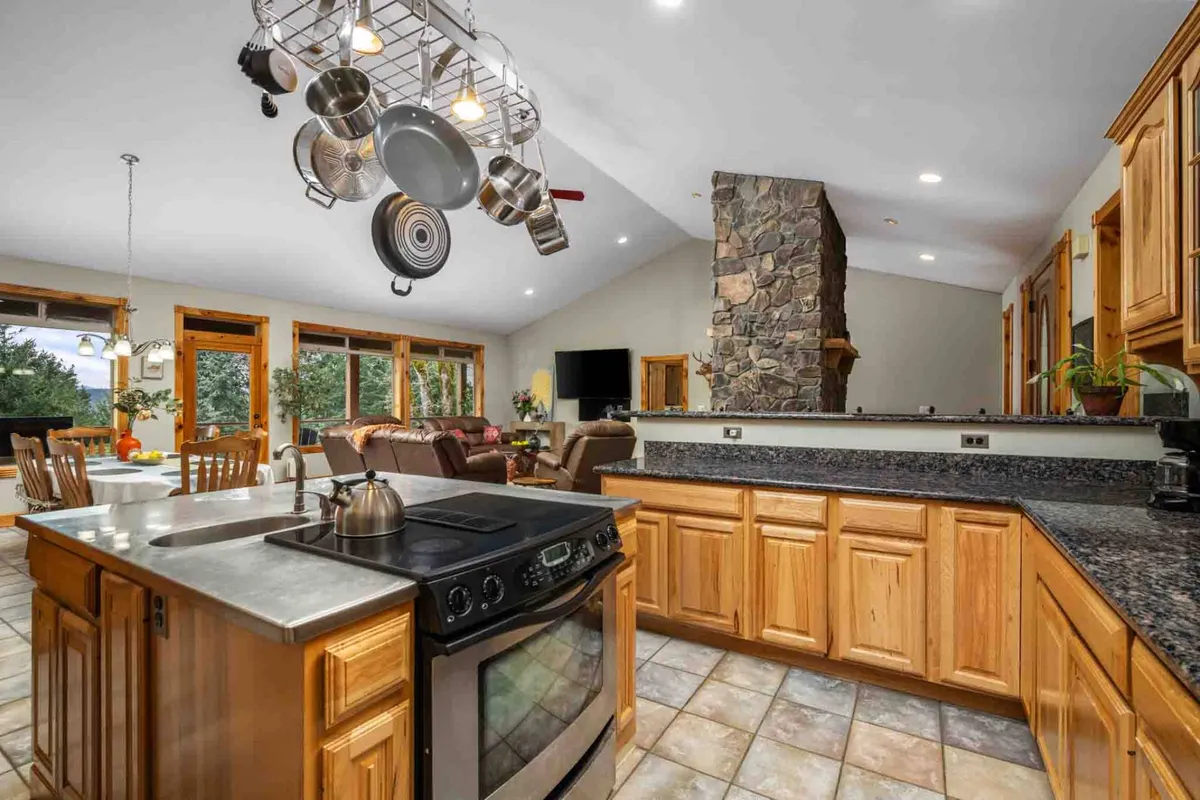 A welcoming open-concept space featuring a prominent stone fireplace, a spacious kitchen island, stainless steel appliances, large windows flooding the area with natural light, and hanging pots that contribute to the lodge-style ambiance. Plush seating and warm wood accents encourage both relaxation and lively gatherings.